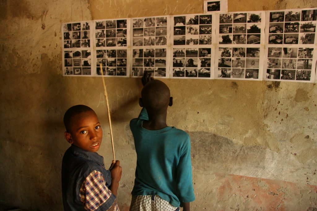 Zwei Jungen unterschiedlichen Alters stehen vor einer Wand und betrachten Abzüge von Fotos, die sie für ein Medienprojekt selbst aufgenommen haben. Uno dei bambini blocca uno zeigestock nella mano e si scarica nella fotocamera.
